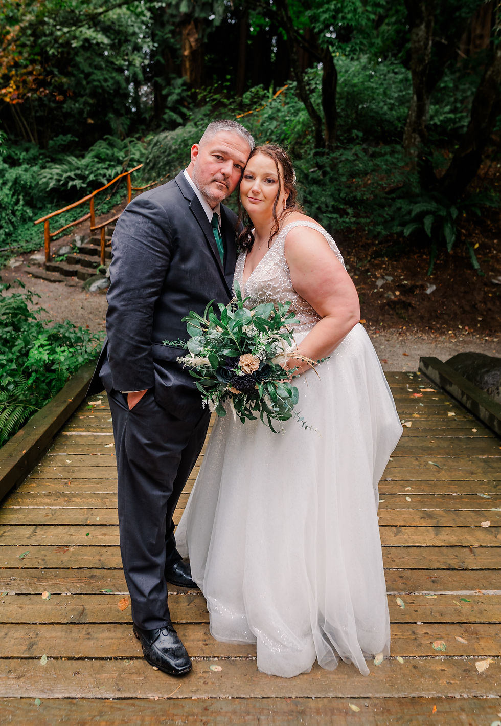 Bride in white gown and groom in dark suit stand on a wooden bridge, surrounded by lush greenery. Bride holds a leafy bouquet. Serene mood.