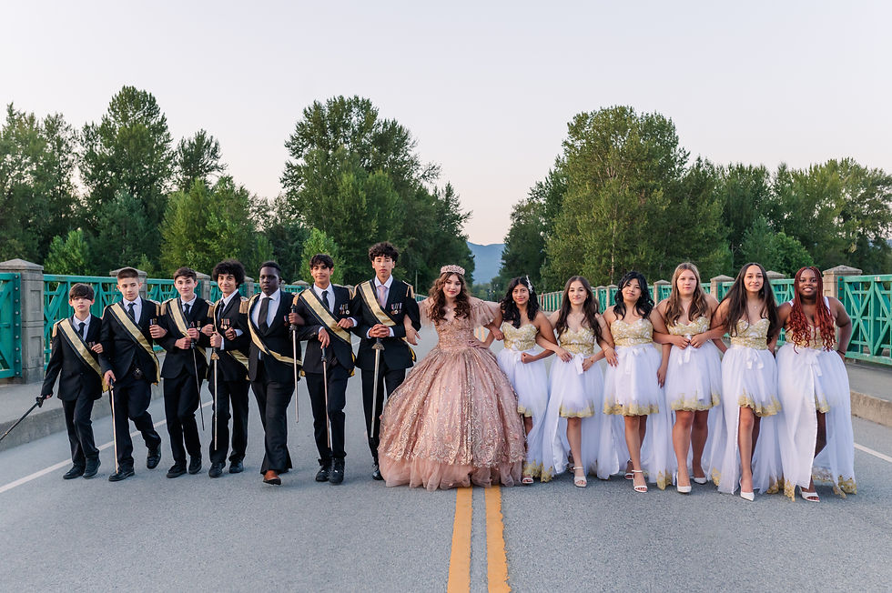 A group of teens in formal attire pose on a road with a forest backdrop. Boys wear black suits, girls in white dresses. Happy and festive mood.