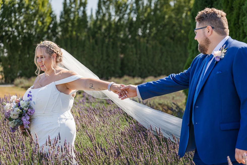 Bride in white dress holding groom's hand in blue suit, standing in a lavender field. Both appear joyful, with trees in the background.
