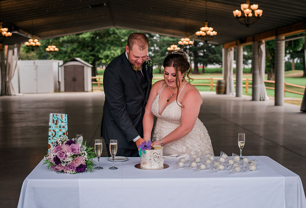 Bride and groom cut cake on a table with purple flowers and champagne, under a pavilion with chandeliers, creating a joyful scene.
