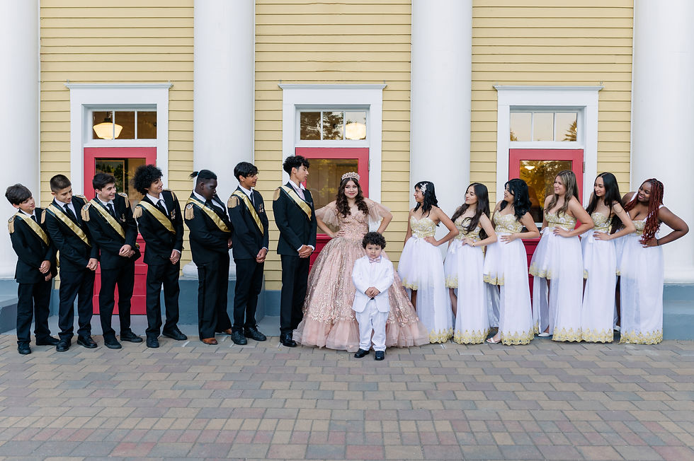 A group photo in front of a yellow and red building. Boys in black suits with gold sashes and girls in white dresses surround a girl in pink. A child stands in front.