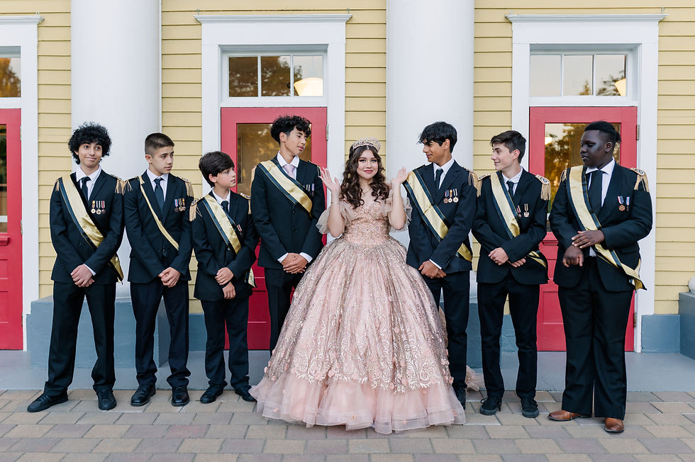 A girl in a pink gown stands smiling with six boys in suits with sashes. They are in front of a yellow building with red doors.