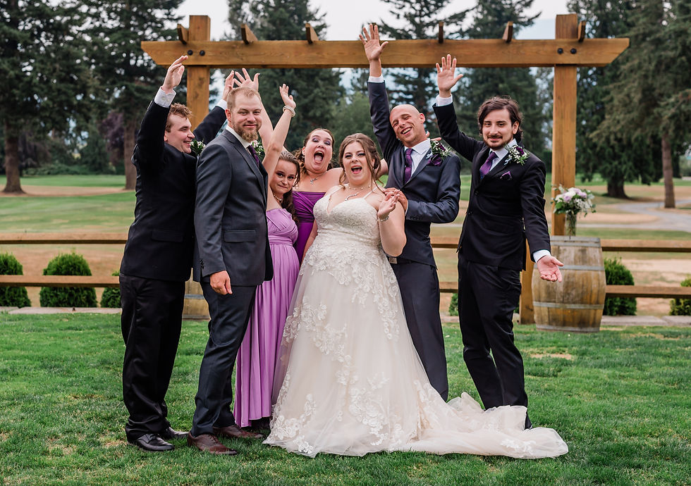 Bridal party cheers joyfully, posing on grass under wooden arch. Bride in white, bridesmaids in purple, groomsmen in dark suits.