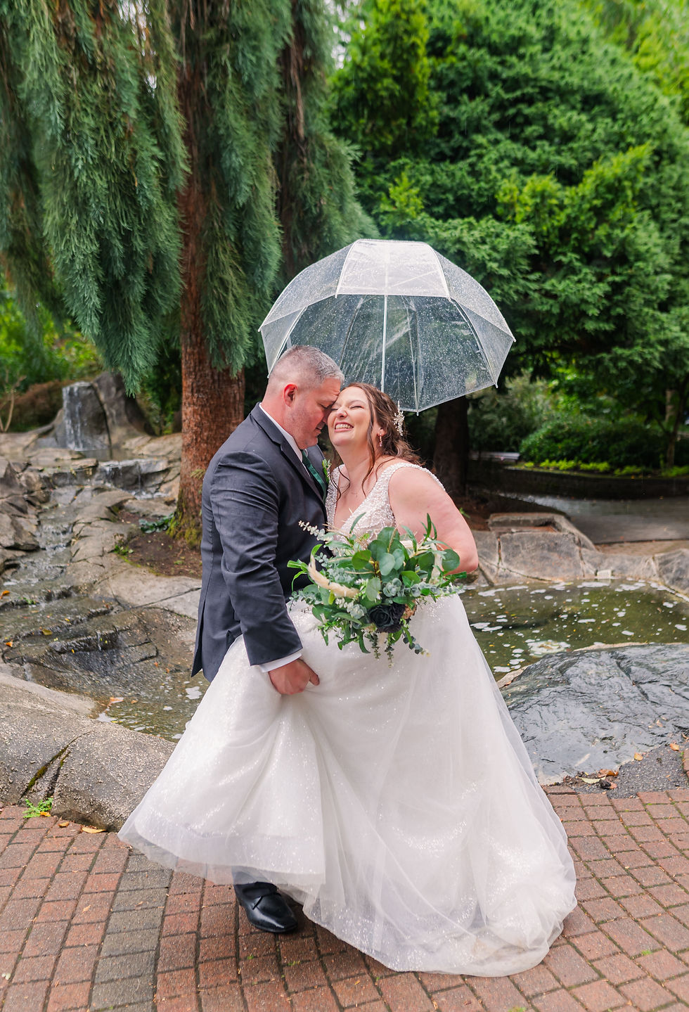 Bride and groom share a joyful moment under a clear umbrella. She's in a white dress holding flowers, surrounded by a lush garden setting.
