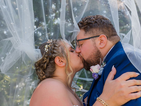 de and groom kissing under a sheer veil with floral details, groom in blue suit. Emotional, romantic outdoor setting.