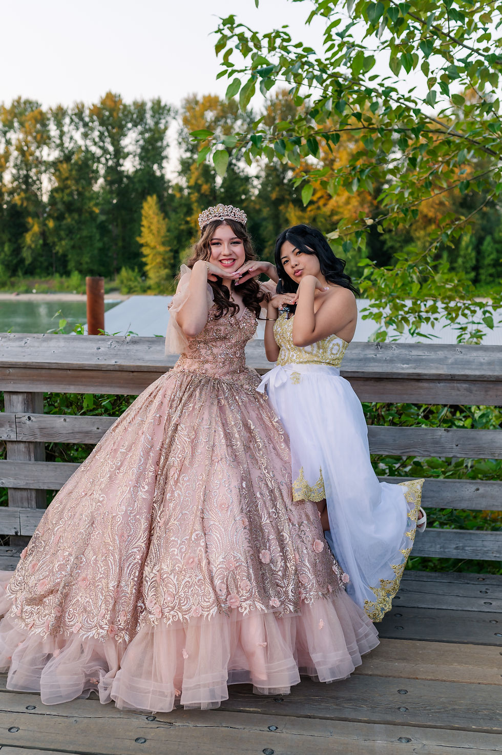 Two women pose playfully by a wooden fence in elegant dresses and crowns. Background has lush greenery and a river, creating a cheerful mood.