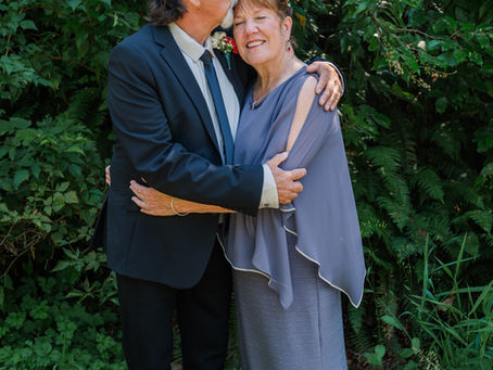 An elderly couple embraces joyfully; the man kisses the woman's forehead. They're outdoors, surrounded by lush greenery. A bouquet lies nearby.