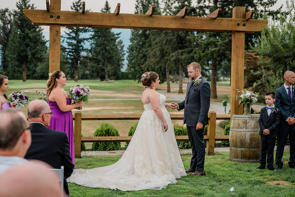 Bride and groom exchange vows under a wooden arch outdoors. Bridesmaids in purple hold flowers. Guests seated. Trees in the background.