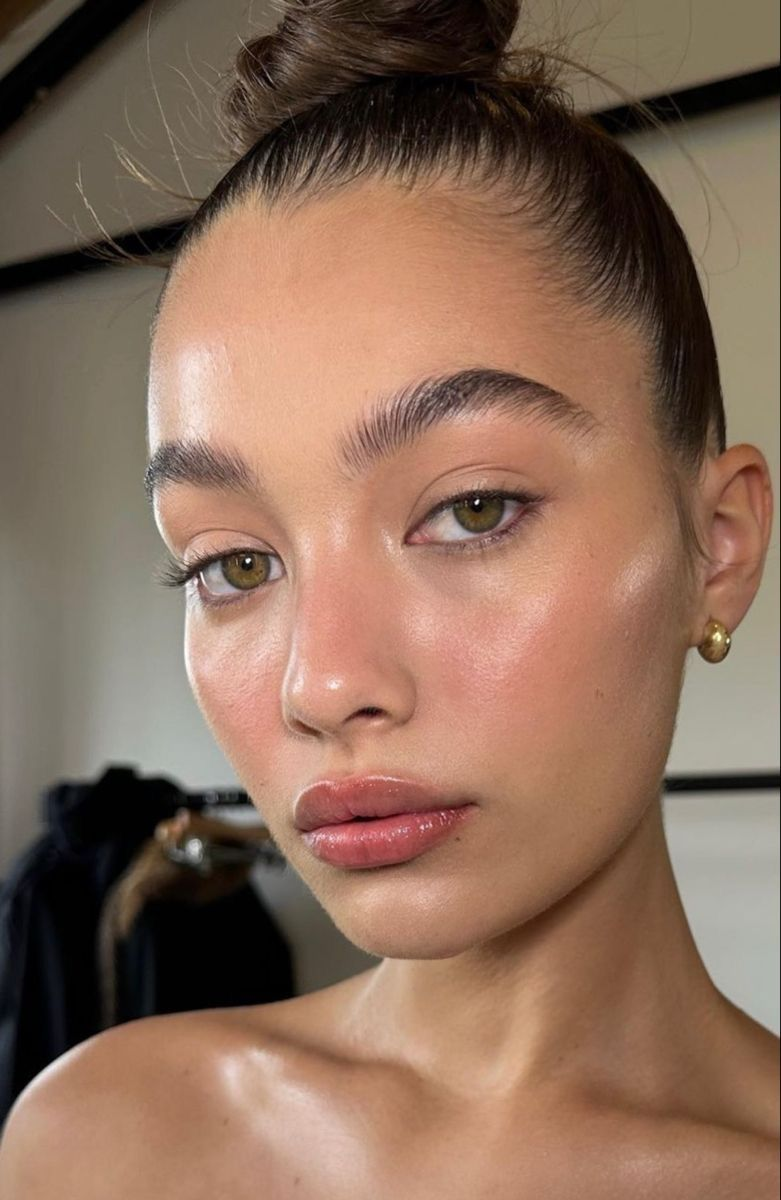 Woman with slick bun and gold earring gazes at camera, with soft natural makeup. Background shows blurred clothes, creating a calm mood.