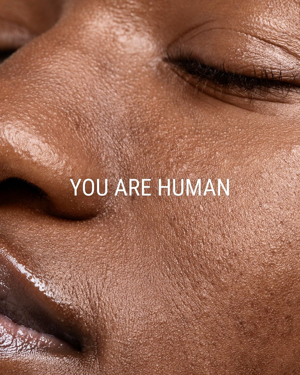 Close-up of a person's peaceful face with closed eyes, smooth brown skin. Text reads "YOU ARE HUMAN" in white letters.