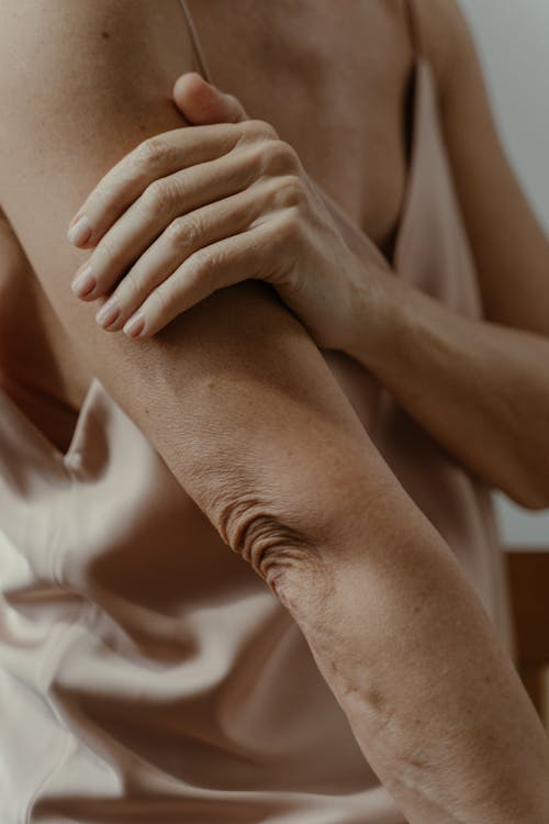 Close-up of a person in a silky, beige top touching their arm. The image emphasizes skin texture and a calm, introspective mood.