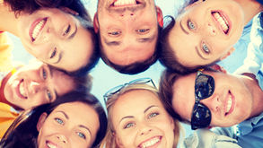 Seis amigos sorridentes em círculo, vistos de baixo, sob céu azul, em clima alegre.