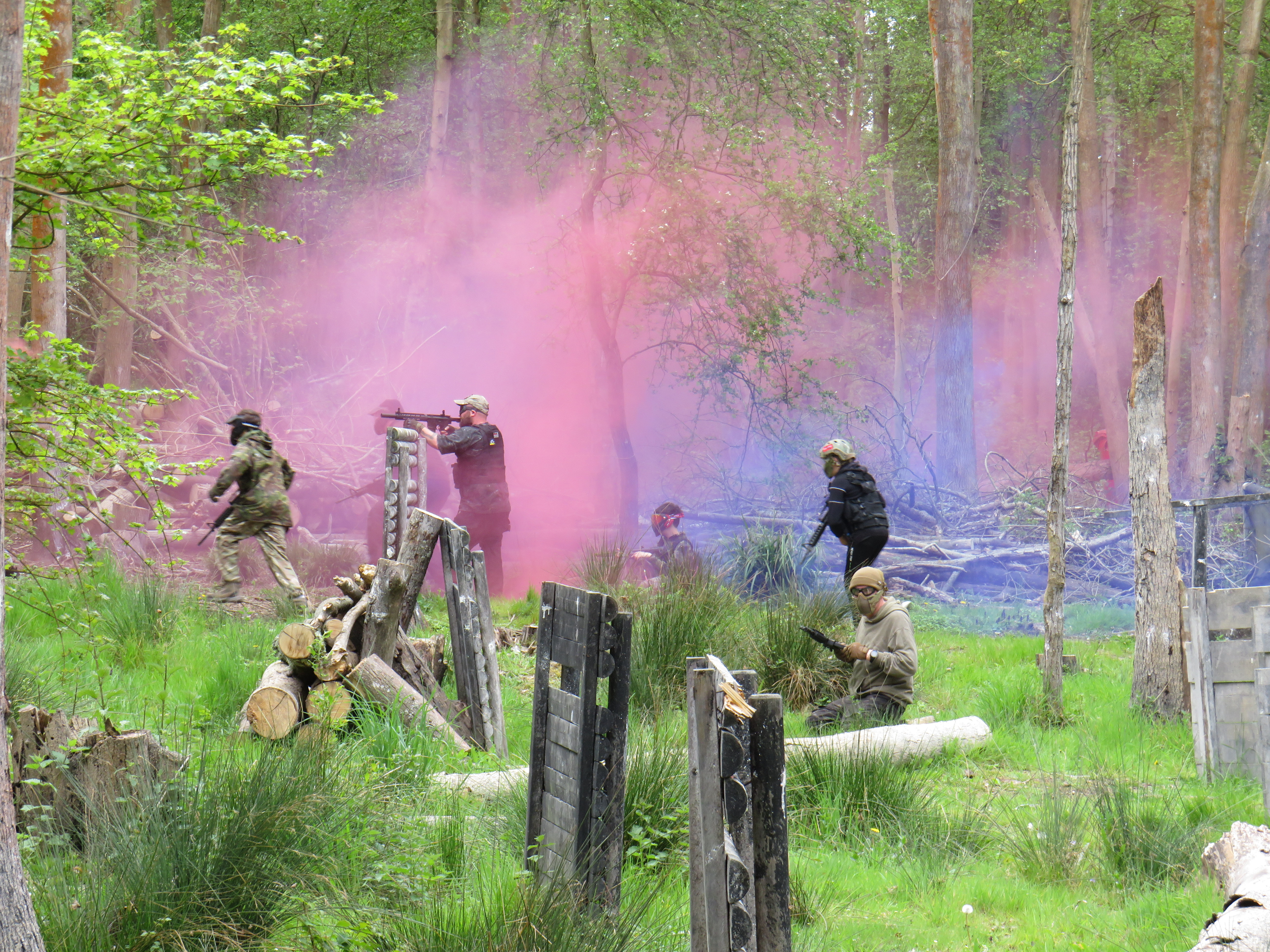 Airsoft players move through a woodland game while pink and blue smoke drifts between the trees.