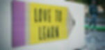 Yellow pencil-shaped sign with "LOVE TO LEARN" text on a wall. A person walking in the blurry background, outdoor setting, casual vibe.