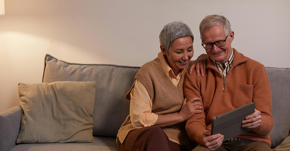 Senior couple smiling and sitting on a couch, sharing a moment while looking at a tablet. Cozy setting with a light brown palette.