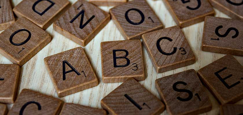 Wooden Scrabble tiles scattered on a light wood surface, featuring letters like A, B, C with numbers. The mood is casual and playful.
