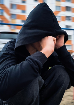 Trauriger-Teenager-sitzt-niedergeschlagen-vor-einem-Haus-er-hockt-in-sich-gekauert-und hat-die-Kapuze-des-schwaren-Hoodie-über-den-Kopf-gezogen-seine-Hände-verstecken-das-Gesicht-er-nimmt-die-Umgebung-nicht-wahr-hat-sich-zurück-gezogen-wirkt-kraftlos-und-depressiv-enttäuscht-verzweifelt