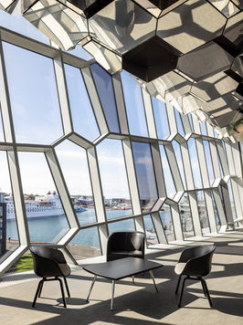 <img src=”- Modern-Reykjavik.jpg” alt=”Contemporary lounge with floor-to-ceiling geometric hexagonal windows overlooking a harbor with boats and waterfront buildings. Three black chairs and a small square table sit in the foreground, illuminated by strong natural light casting angular shadows across the floor. The honeycomb-patterned glass façade and sculptural ceiling elements emphasize the modern architectural design.”/>