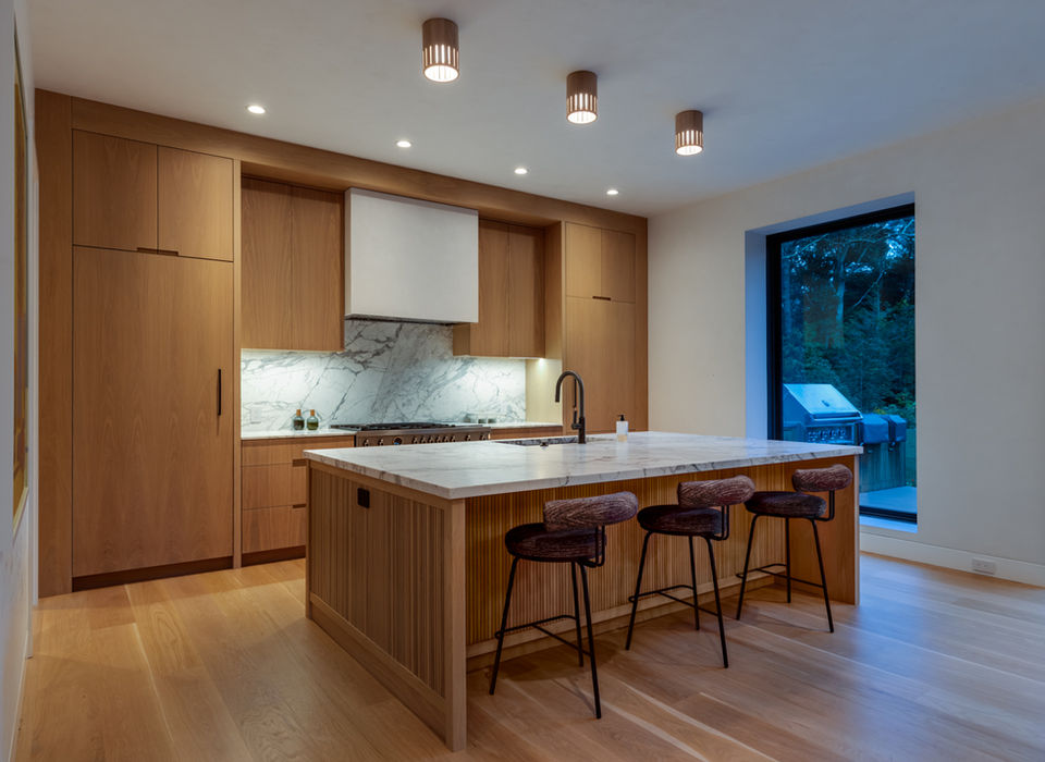 <img src=”modern-kitchen-Amagansett.jpg” alt=”A sleek modern kitchen with warm wood cabinetry, marble backsplash, and matching marble countertops. A large central island with vertical wood paneling and three upholstered barstools provides seating and workspace. Recessed ceiling lights and three pendant fixtures illuminate the area, while a large window offers a view of an outdoor grill and greenery. The space combines natural materials and clean lines for a contemporary, inviting look”/>