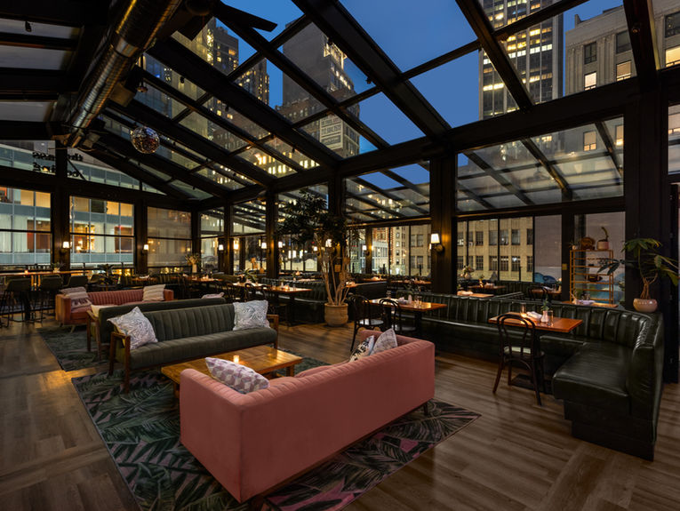 <img src="Elegant - contemporary - rooftop bar restaurant - New York City.jpg" alt="Spacious glass-enclosed rooftop lounge at dusk with city skyline views, featuring booth seating, sofas, wooden tables, and warm ambient lighting."/>