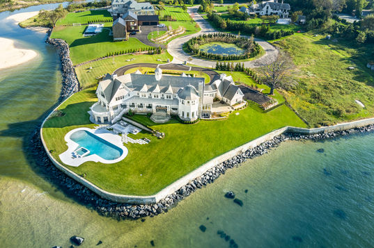 <img src=”Sprawling -Traditional -Estate -Sands Point.jpg” alt=”Aerial view of a luxurious coastal estate with turret-style architecture, a large curving pool with attached hot tub, manicured lawns, and a stone seawall lining the water's edge”/>