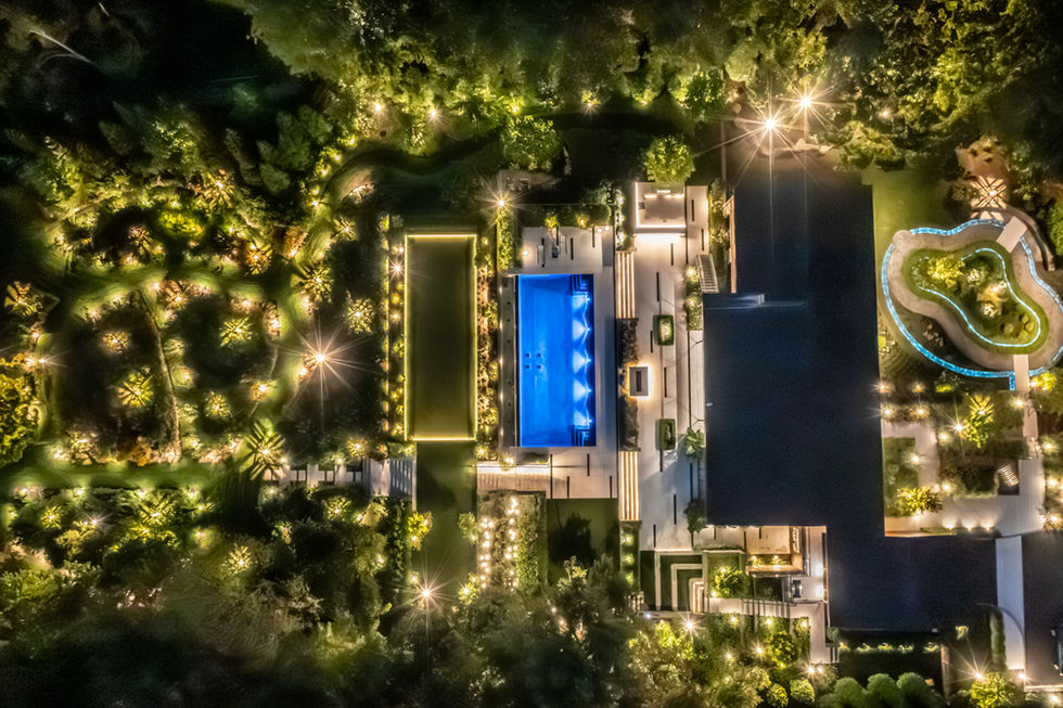 <img src=”Luxurious-contemporary-house-Sands Point.jpg” alt=”Stunning aerial night view of a lavish estate with a glowing blue rectangular pool, formal gardens, and surrounding pathways lit by landscape lighting throughout the property”/>