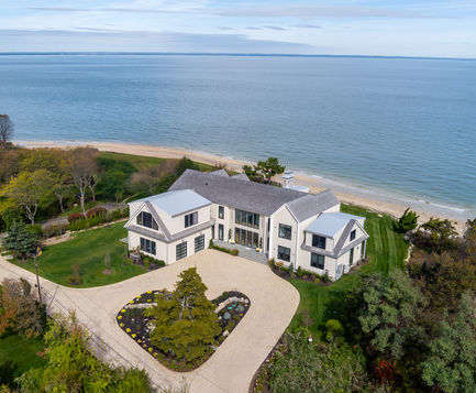 <img src=”Sprawling -Contempary Home -Estate - Fort Salonga.jpg” alt=”An aerial view of a large modern coastal home situated along a sandy beachfront. The house features light-colored exterior walls, gabled roofs, and expansive windows overlooking the ocean. A circular driveway with landscaped greenery leads to the front entrance, surrounded by manicured lawns and mature trees. The serene blue water and shoreline create a picturesque backdrop for the elegant seaside property”/>