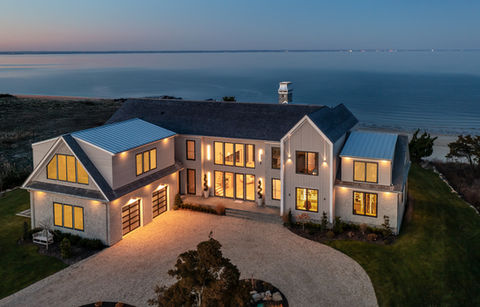 <img src=”coastal-modern-house- Lloyd Harbor.jpg” alt=”Coastal modern home at dusk overlooking the water, featuring large windows, a gravel driveway, and warm interior lighting.”/>