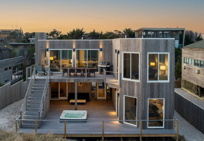 <img src=”fancy-contemporary-house-Fire island.jpg” alt=”Modern two-story beach house with a weathered wood exterior, large windows, and a wraparound deck featuring a hot tub, outdoor seating, and a barbecue grill, all illuminated by soft evening lighting”/>