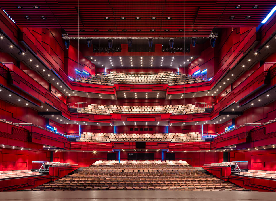 <img src=”Modern-Theater interior-Reykjavik.jpg” alt=”A grand, modern concert hall with multiple tiers of seating arranged in a semi-circular layout. The interior is dominated by deep red walls and black accents, with rows of beige seats illuminated by spotlights and blue ambient lighting. The stage is at the bottom of the image, viewed from the performer's perspective, facing the audience seating that rises across several balconies”/>