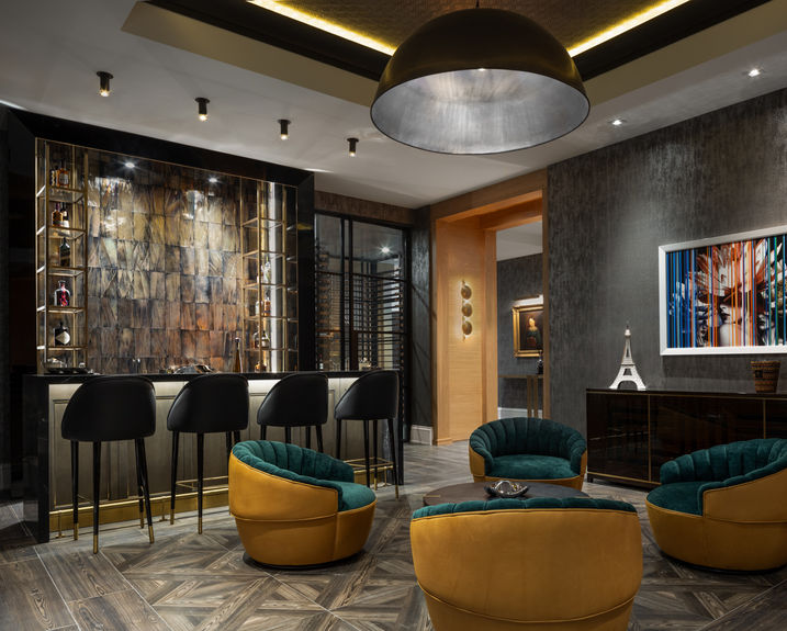 <img src="Luxury - contemporary - home - Upper Brookville.jpg" alt="Modern lounge with a small bar, black stools, and illuminated bottle display, featuring curved velvet chairs, patterned flooring, and warm, upscale interior lighting."/>
