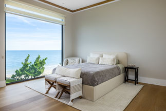 <img src=”elegant-contemporary-bedroom-Fort Salonga.jpg” alt=”A serene bedroom with a large floor-to-ceiling window offering a stunning ocean view. The room features a neutral color palette with light wood floors, beige walls, and a cream upholstered bed dressed in gray and white linens. Two cushioned benches with throws sit at the foot of the bed, and a small side table with decorative accents stands beside it. Natural light fills the space, creating a calm and airy coastal atmosphere overlooking the Long Island Sound”/>
