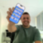 Man in a green shirt smiles, holding a smartphone displaying colorful app icons. Bright indoor setting with white walls and a black chair.