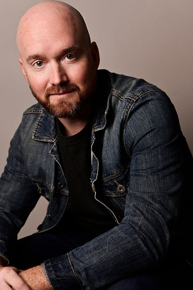 Ned Donovan's headshot of a neutral face looking at the camera while sitting and his hands folded on his knees while wearing a black t-shirt and a jean jacket.