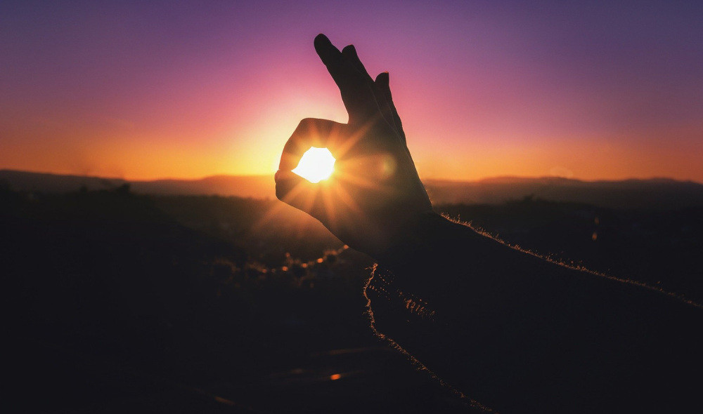 Hand making 'okay' sign in front of sunset