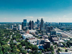 Aerial view of the Atlanta skyline showing Midtown skyscrapers, surrounding neighborhoods, and a major highway corridor cutting through the city under a clear blue sky.