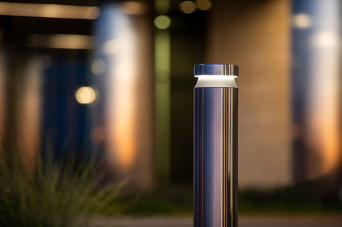 a close-up of a stainless steel bollard with a light ring at the top, set in an outdoor environment with a blurred background.
