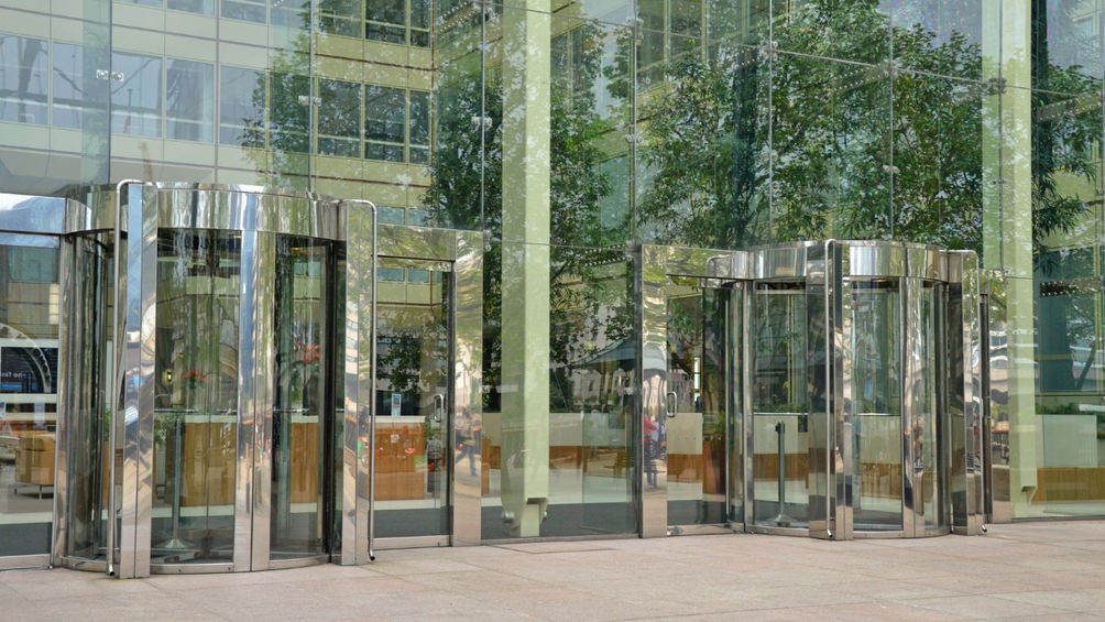 a set of revolving doors made of stainless steel, set in front of a glass building with reflections of trees and the surrounding environment.