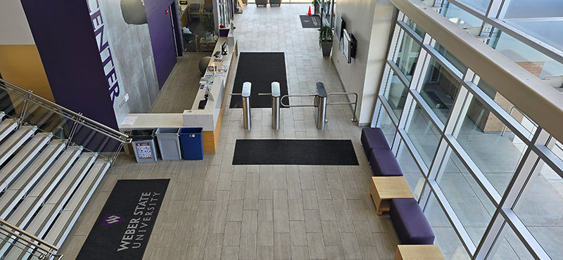 overhead view of a university lobby with waist-height turnstiles and benches, with "Weber State University" logo visible on the floor mat.