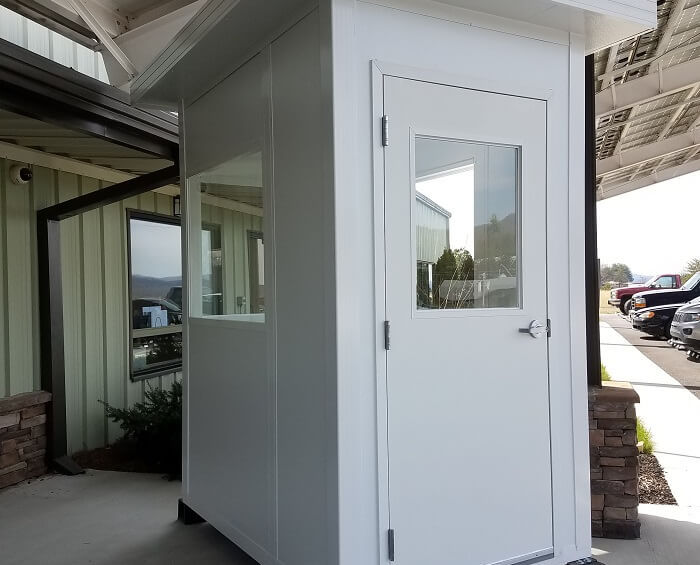 small white security booth with a door and a window, located outside near a building with vehicles in the background.