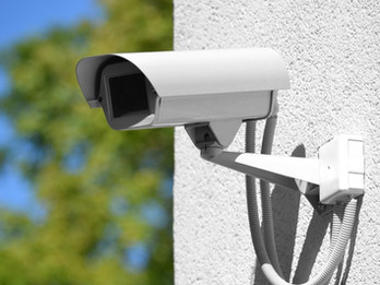 a white security camera mounted on a wall with visible wiring and a blurred background of greenery.