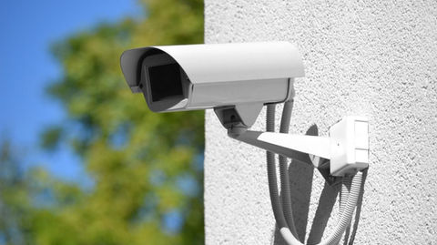 a white security camera mounted on a wall with visible wiring and a blurred background of greenery.