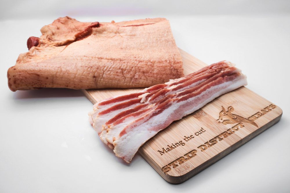Half slab of bacon, with a few fresh cut slices on a strip district meats cutting board.