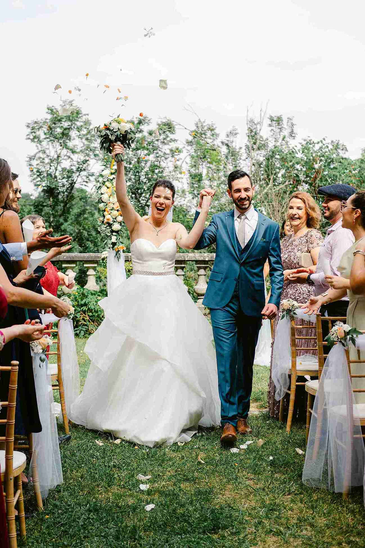 deux mariés s'avancent dans une allée en extérieur entourés de leurs proches. La mariée pousse un cri de joie, son bouquet à la maison. Elle à une longue robe blanche et lui porte un costume bleu marine