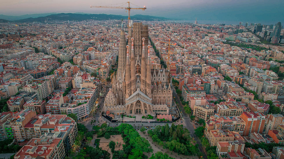 Aerial view of Sagrada Familia in Barcelona, surrounded by buildings. A crane is visible atop the basilica. Cityscape extends to distant hills.