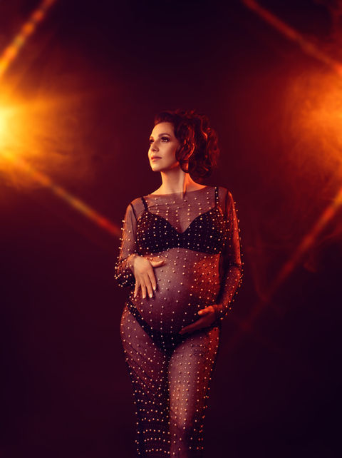 Femme enceinte en body transparent orné de perles, posant sous un éclairage doré et chaleureux, créant une ambiance glamour et puissante.