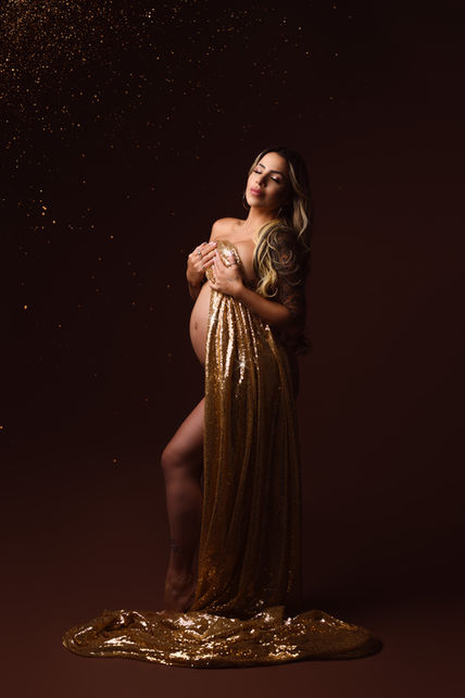 Femme enceinte en robe dorée scintillante, posant sous un éclairage tamisé avec des reflets lumineux au sol, capturant l’élégance et la puissance de la maternité.