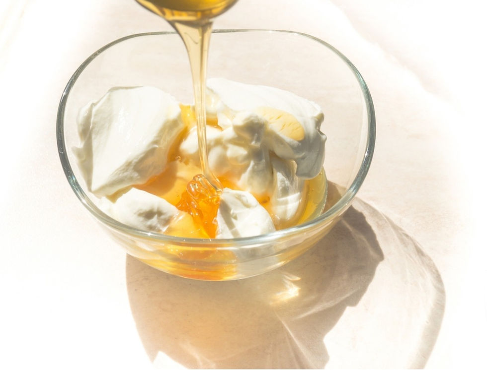 Honey pours over Greek yogurt and in a glass bowl, illuminated by soft sunlight on a light-colored surface.