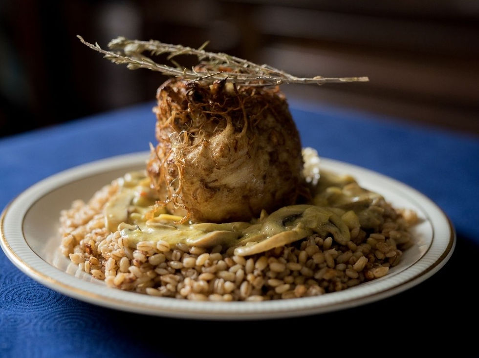 Gourmet dish with a roasted celeraic on a bed of grains, topped with herbs and sauce, on a white plate with blue tablecloth. Warm light.