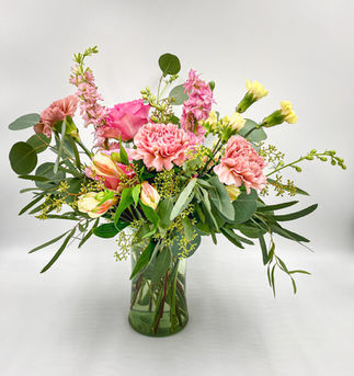 Loose, airy arrangement of light pink roses, carnations, larkspur, pale yellow alstroemeria and mini carnations, eucalyptus, and greenery in a clear glass vase.